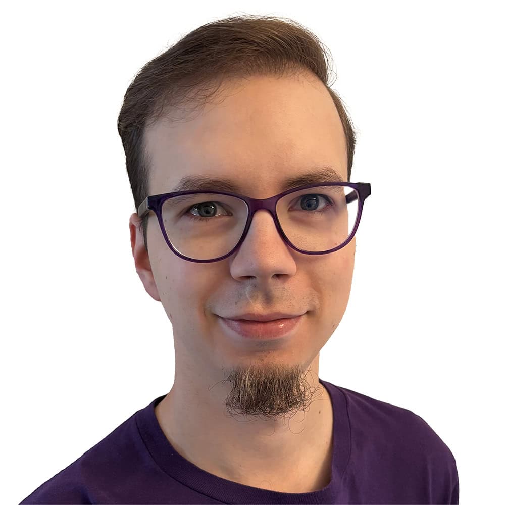 Basti mit einer violetten Brille, violettem Pullover und Bärtchen am Kinn. Er lächelt in die Kamera und steht vor einem weißen Hintergrund.