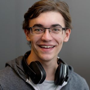 Das Bild zeigt ein Porträt eines jungen Mannes mit braunen Haaren und einer Brille. Er trägt einen grauen Hoodie und um seinen Hals liegen schwarze Kopfhörer. Er lächelt freundlich in die Kamera. Der Hintergrund ist einfarbig grau.  Das Bild wirkt insgesamt positiv und freundlich.